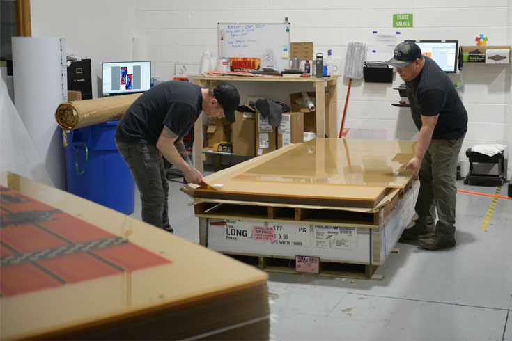 Two technicians carefully inspecting and preparing a large-format rigid print on a production table.